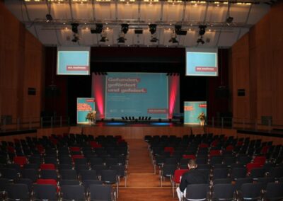 Aula mit leeren Stühlen und einer auf der Bühne bereits laufenden Präsentation