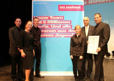 Zwei Frauen und vier Männer stehen vor einem Plakat von "Das Handwerk"